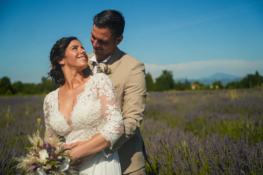 Couple de mariés enlacés dans un champ de lavande, montagnes à l'horizon