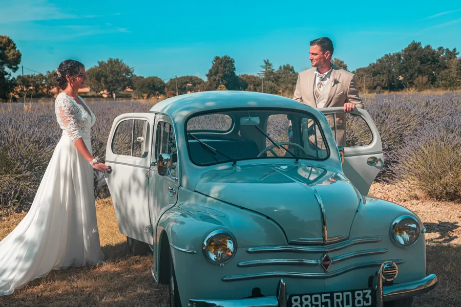 Mariée et marié de part et d'autre d'une voiture ancienne bleu clair, au milieu des lavandes